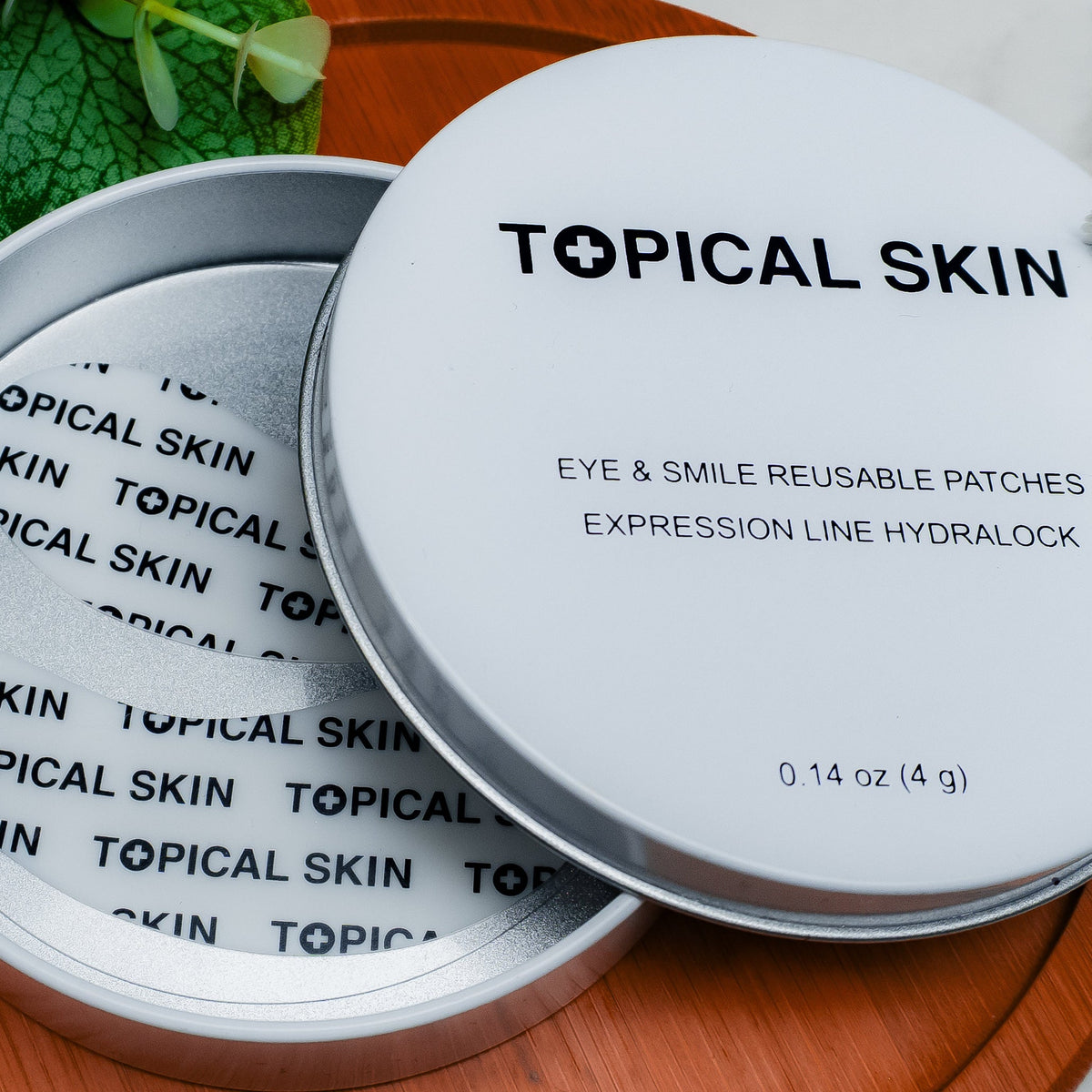 A round silver metal tin of TOPICAL SKIN reusable eye and smile patches, shown open with the lid leaning against the base on a wooden tray with green leaves.