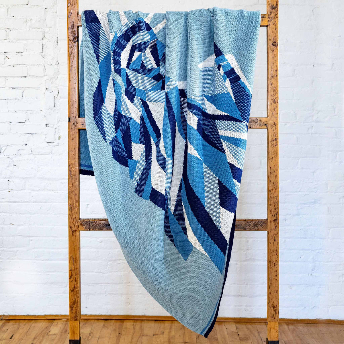 The Statue of Liberty | We the People - Cotton Sustainable Knit Modern Throw Blanket by in2green, featuring a blue and white geometric pattern, is elegantly draped over a wooden rack against a white brick wall and wooden floor—a captivating piece of heritage home decor that infuses any space with timeless charm.