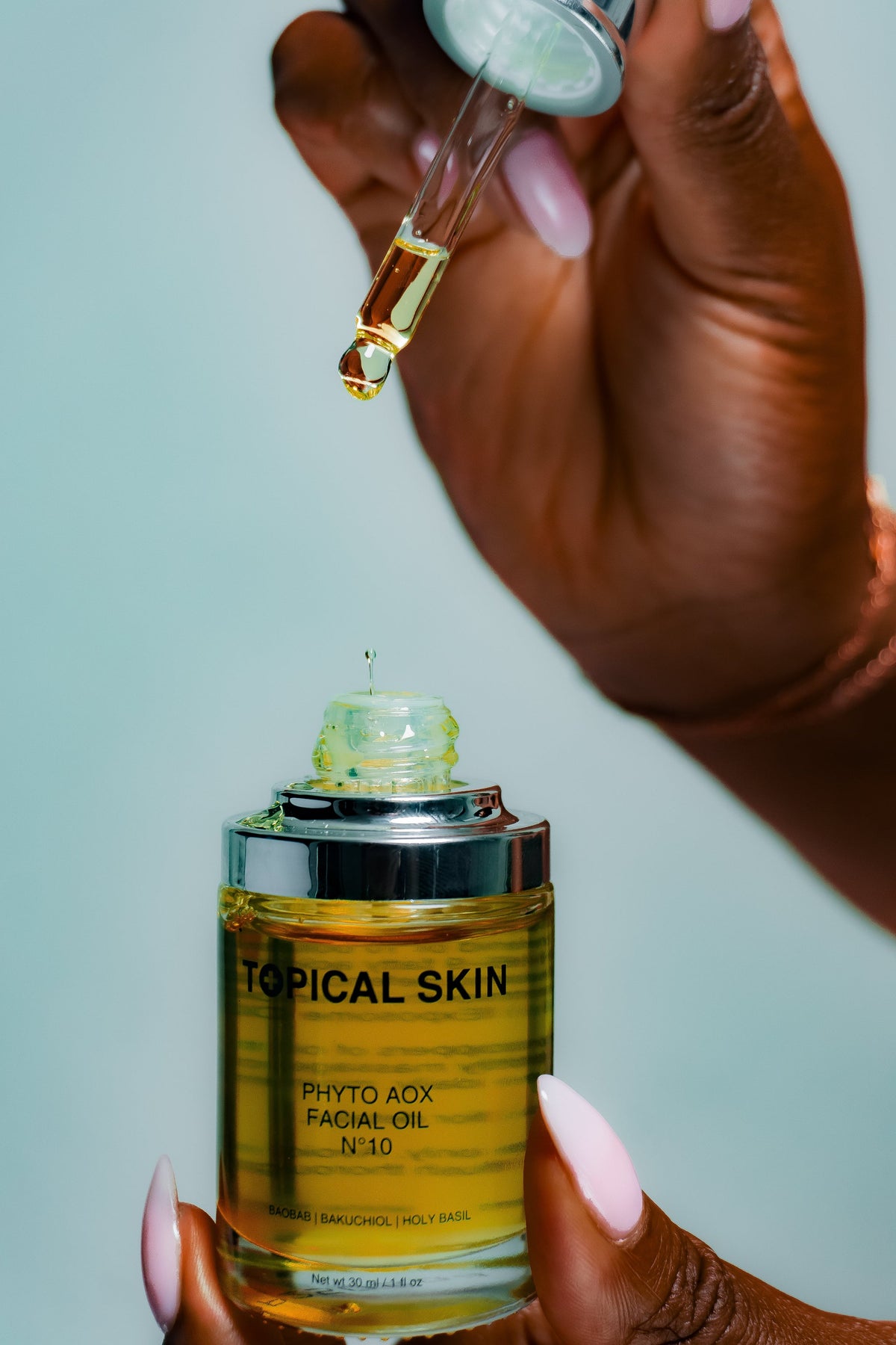 A close-up of a hand holding a clear dropper above a bottle of Topical Skin Phyto Aox Facial Oil N.10, with golden oil dripping from the dropper into the bottle, set against a solid pale blue background.