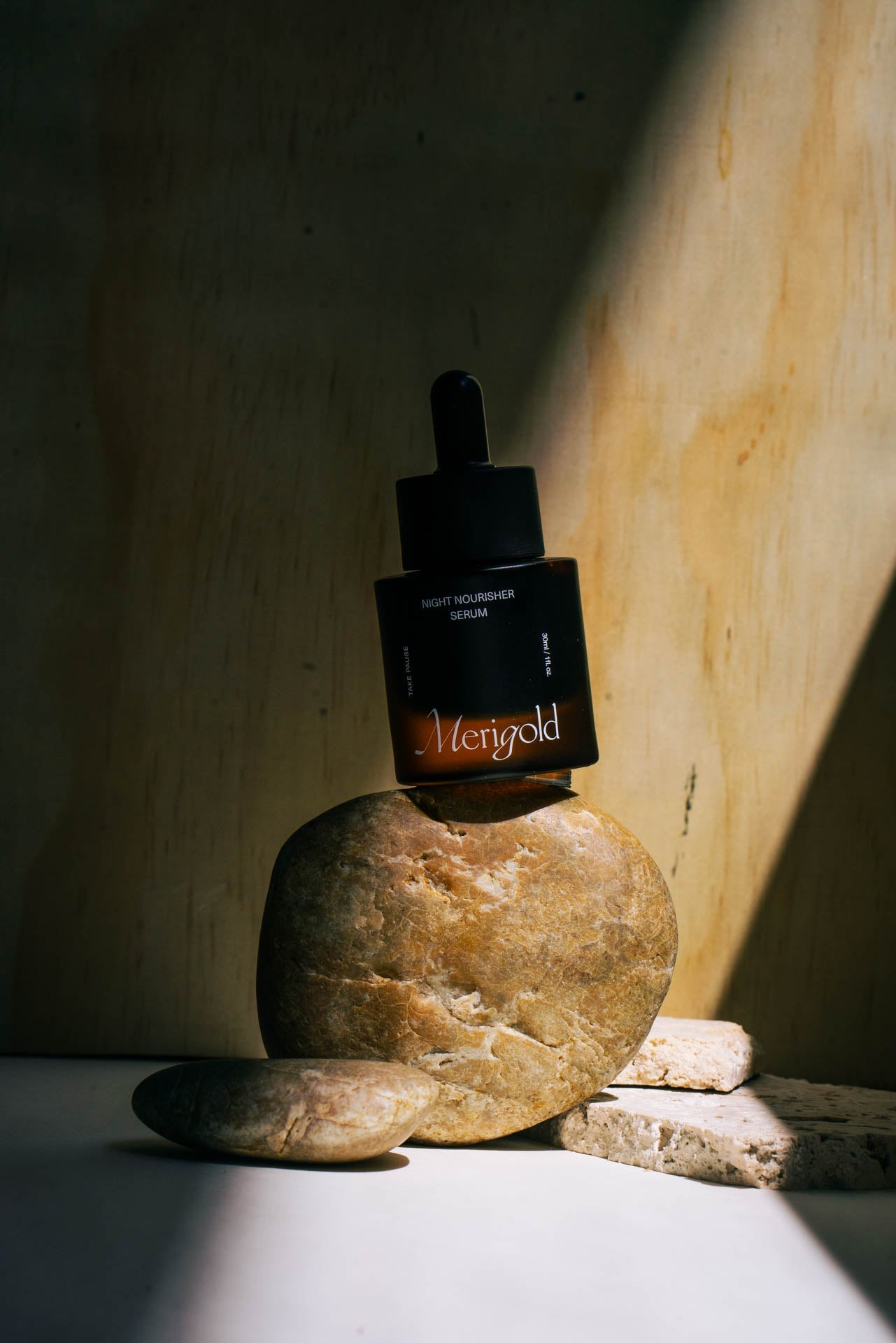 Merigold Night Nourisher Serum bottle balanced on natural stones with dramatic sunlight — premium night facial oil for deep skin nourishment