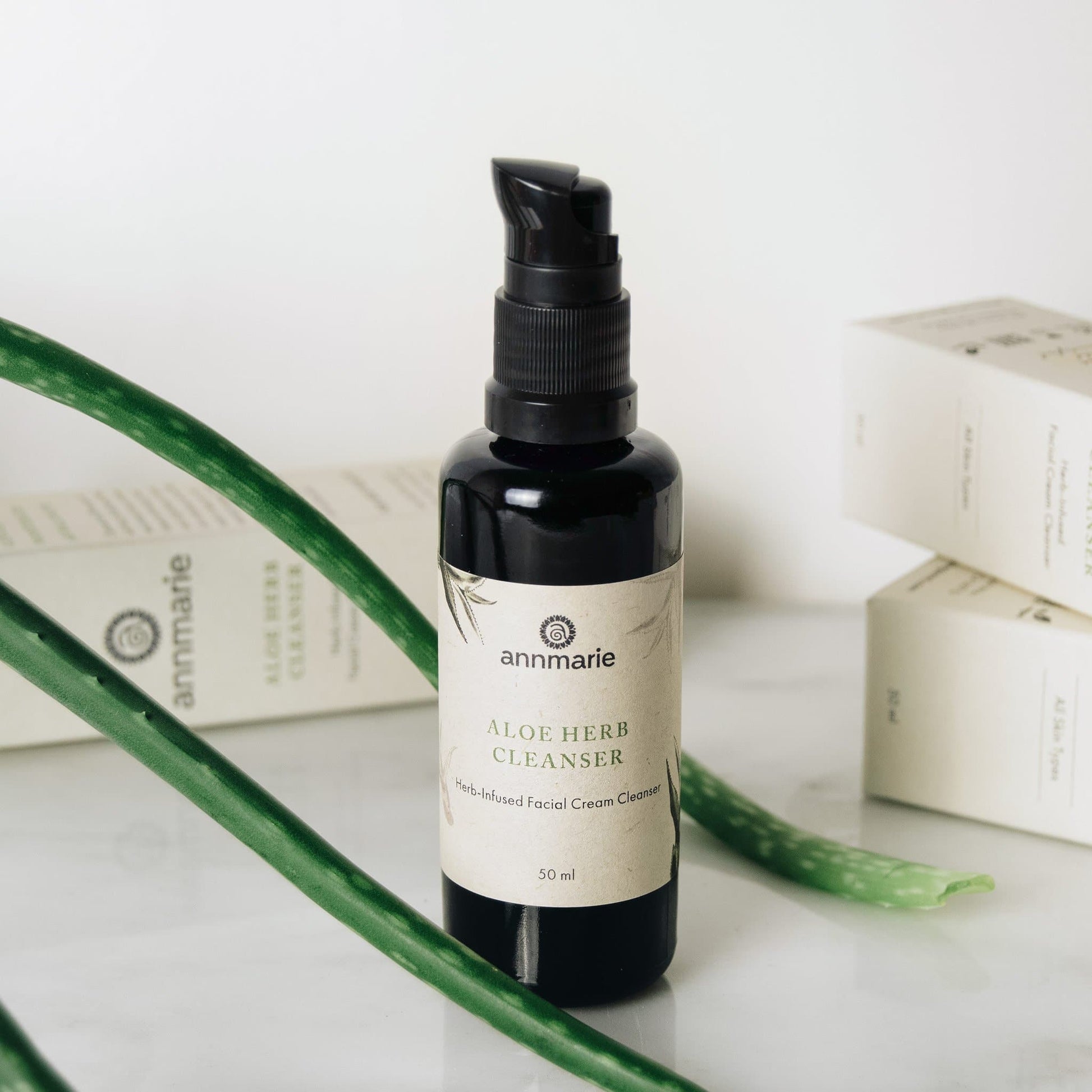 A black pump bottle labeled 'Aloe Herb Cleanser' 50 ml, with the package box and green aloe leaves in the background.
