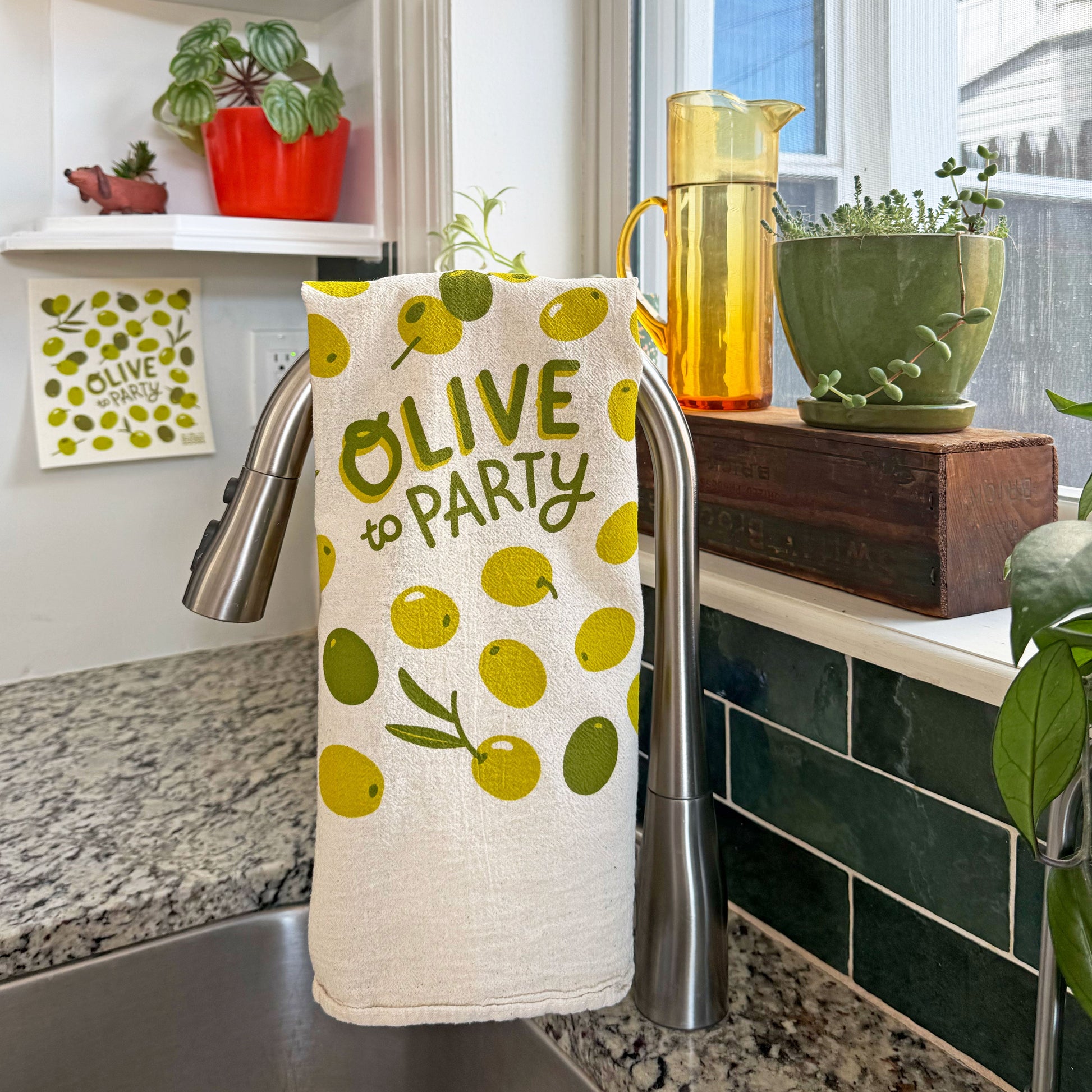 The Neighborgoods Olive dish towel hangs over a kitchen sink faucet and the olive sponge cloth hangs on a wall hook.