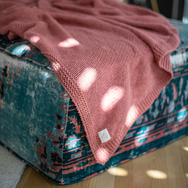 Rosewood pink knitted throw draped over a textured and multi-coloured blue futon.