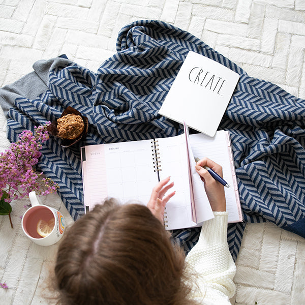 Child sitting on a white rug with a navy blue herringbone patterned blanket, writing in a notebook with a cup of lemonade.