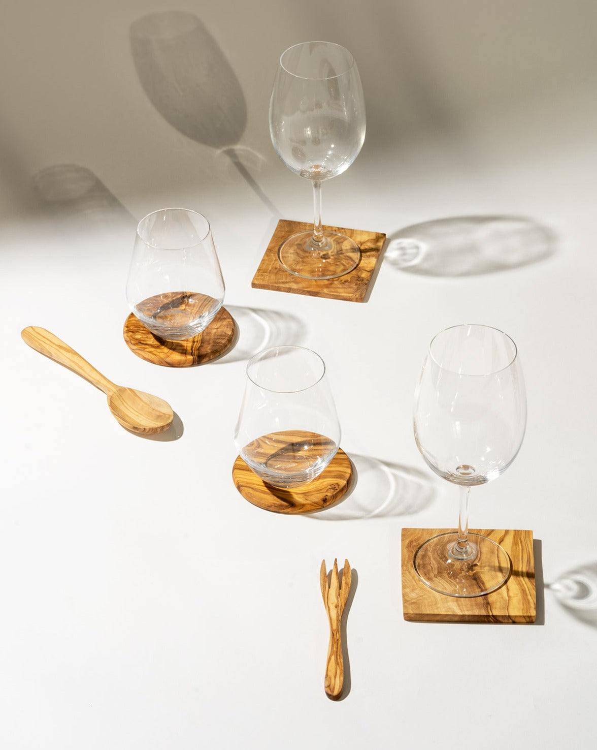 Four square olive wood coasters, each holding a glass, arranged on a white surface. A wooden spoon and fork are placed beside them, showcasing a cohesive, natural tabletop setting.