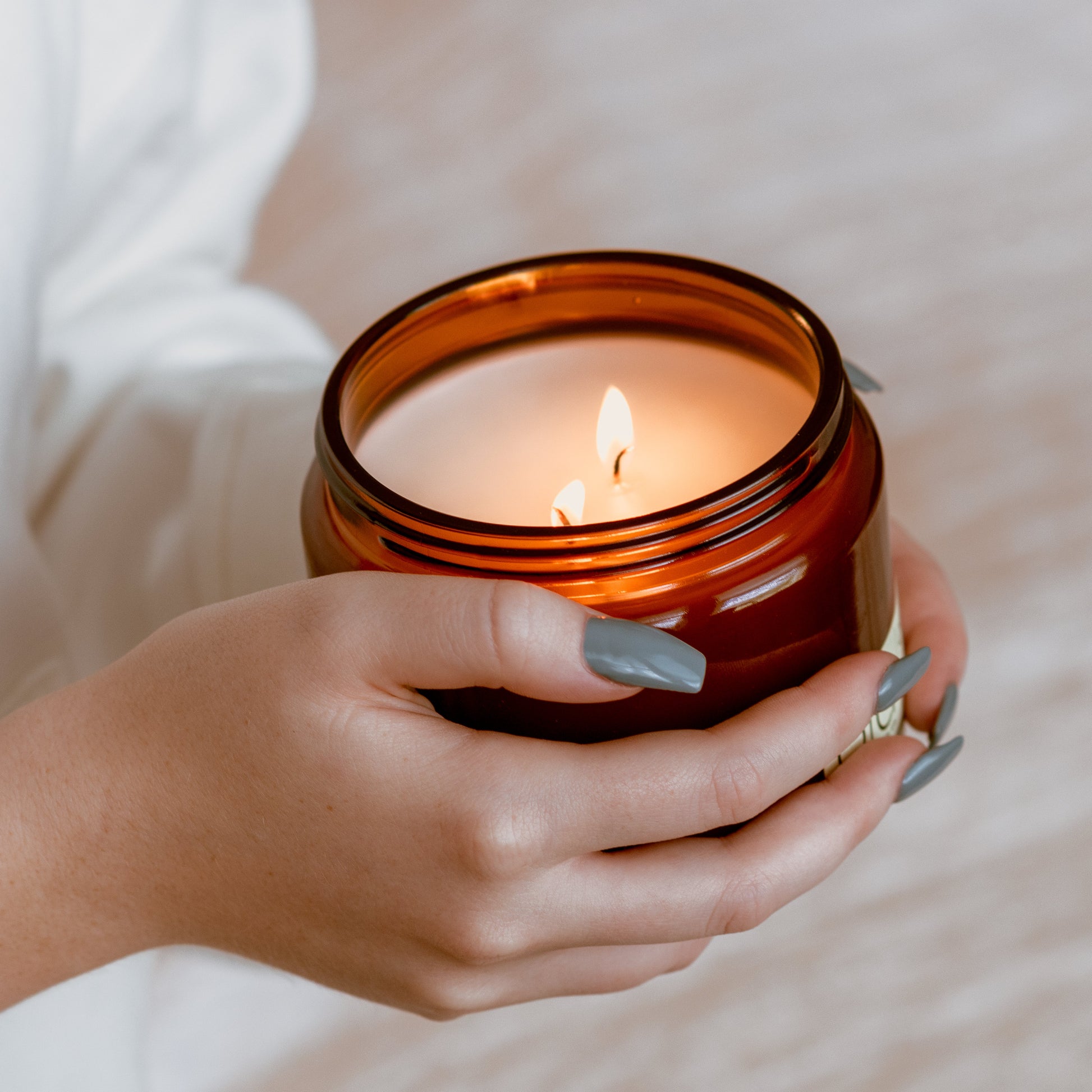 A lit amber glass jar candle held in two hands with gray manicured nails, set against a soft, light beige background.