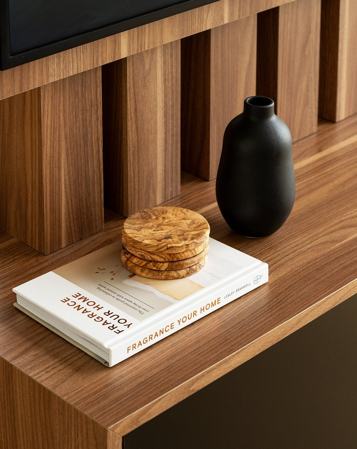Three round olive wood coasters stacked on top of a closed book, placed on a wooden surface. A black empty vase sits beside them, creating a warm and minimal tabletop scene.
