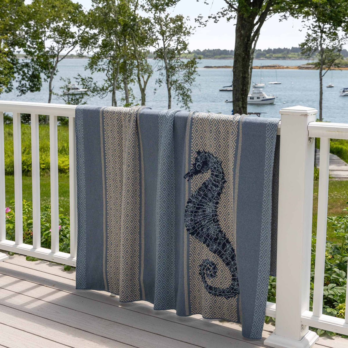 A Seahorse Square - Cotton Sustainable Knit Throw Blanket by In2Green, featuring a coastal blue and beige seahorse design, hangs over a white railing with boats on the water and trees in the background.