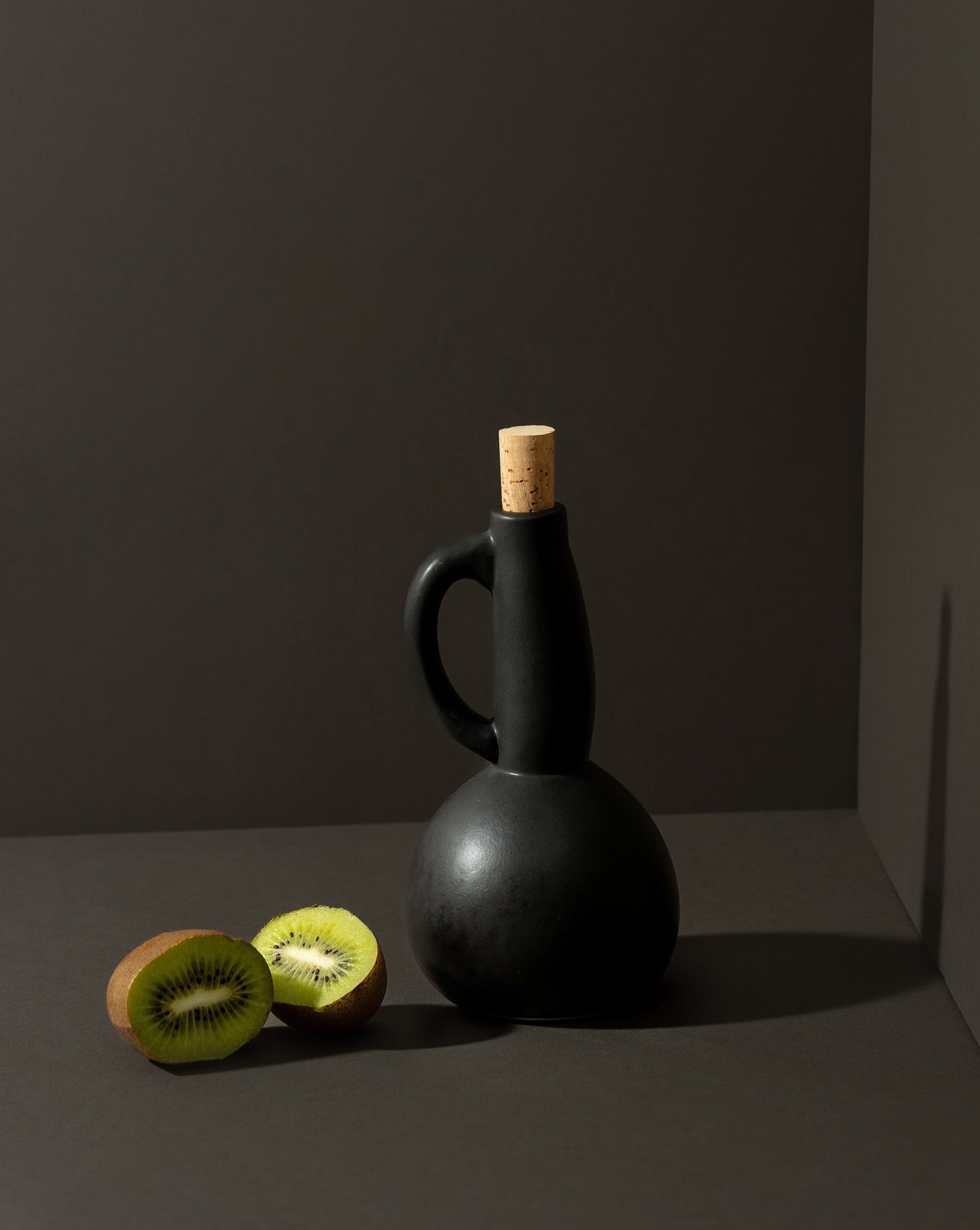 A white ceramic olive oil dispenser with a matte finish, a cork stopper, and a handle, displayed in a corner setting with nuts scattered in front.