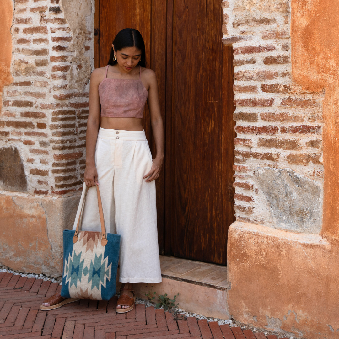 Woman carrying handwoven bucket bag in rose and blue with abstract diamond and lightning pattern, leather shoulder straps, and relaxed shape, ideal for everyday or beach use.