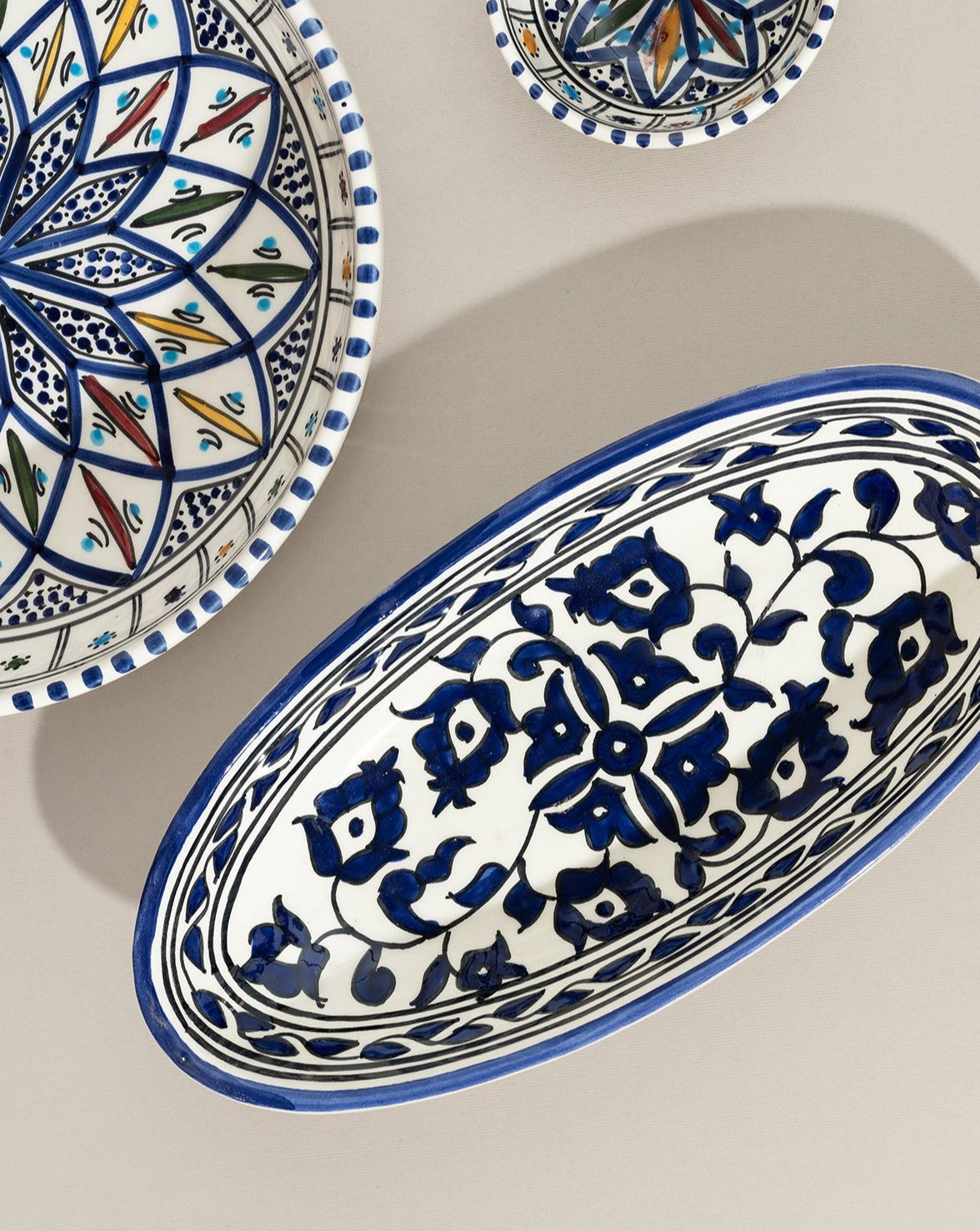 Three hand-painted ceramic dishes on a neutral background—an oval plate with a blue floral design, a large round plate with a colorful geometric pattern, and a small round dish with a star design.
