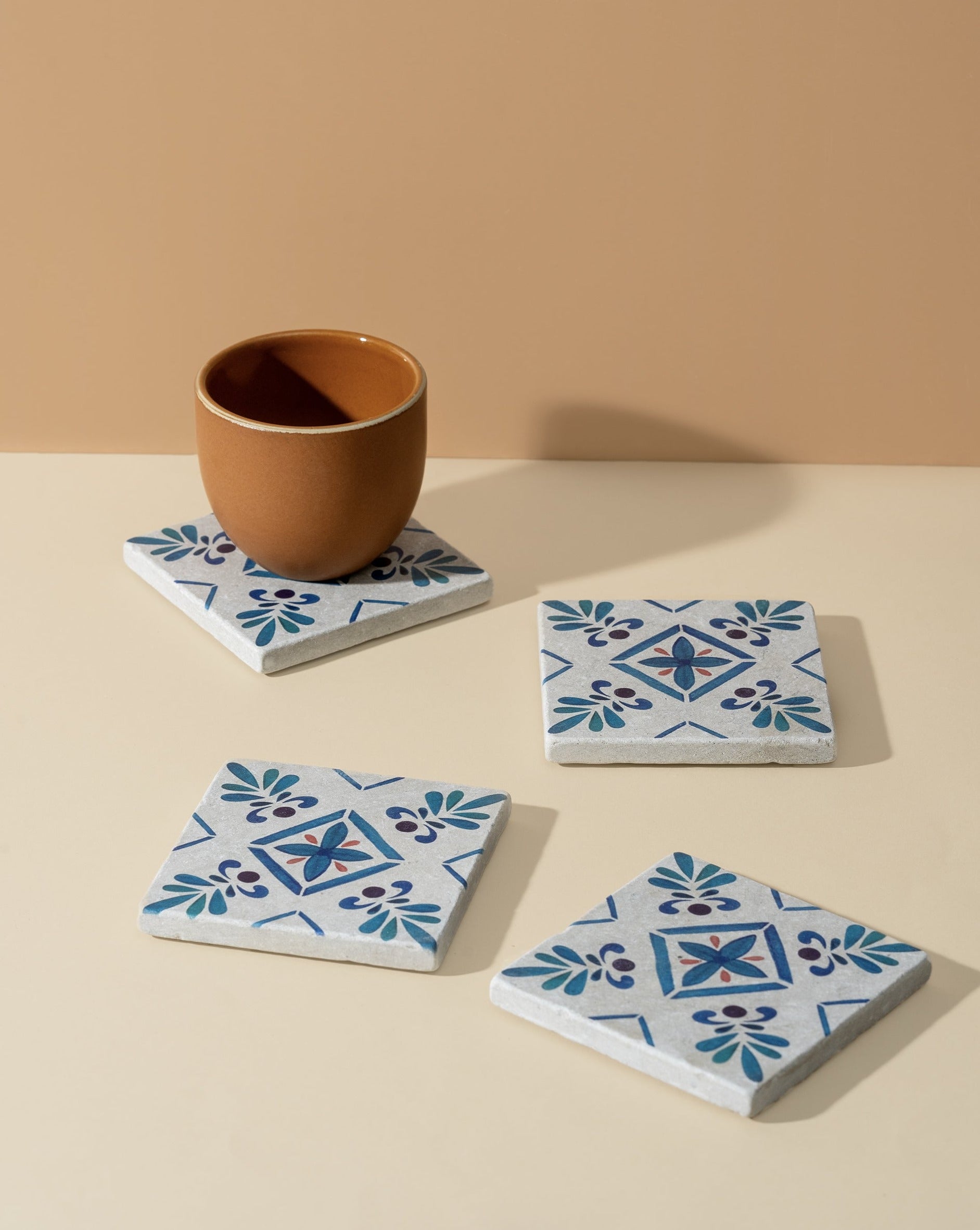 A set of four square white marble coasters with blue floral patterns, arranged on a surface with a cup placed on one of them, highlighting both design and function.
