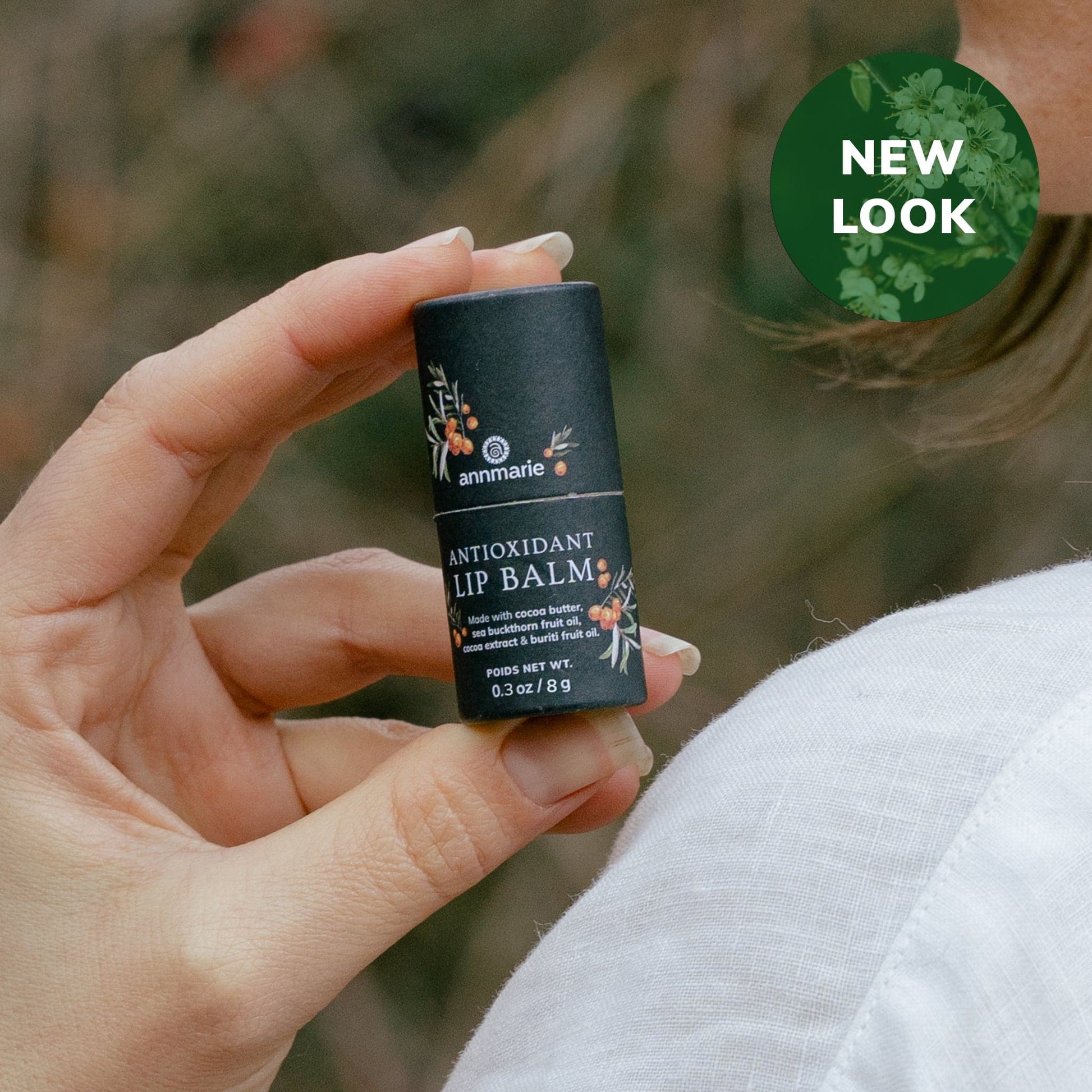 Hand holding Annmarie Skin Care Antioxidant Lip Balm in black tube packaging with botanical design and “New Look” badge in the corner.