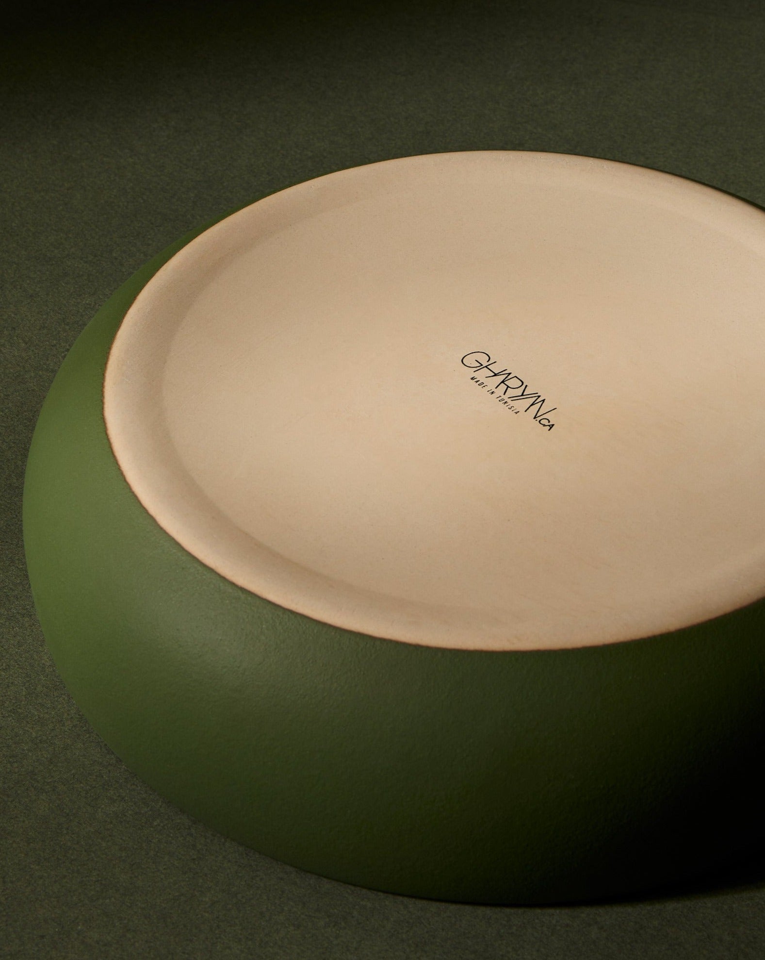A matte green stoneware shallow serving bowl placed upside down against a dark background, showing the “GHARYAN” logo on the base.