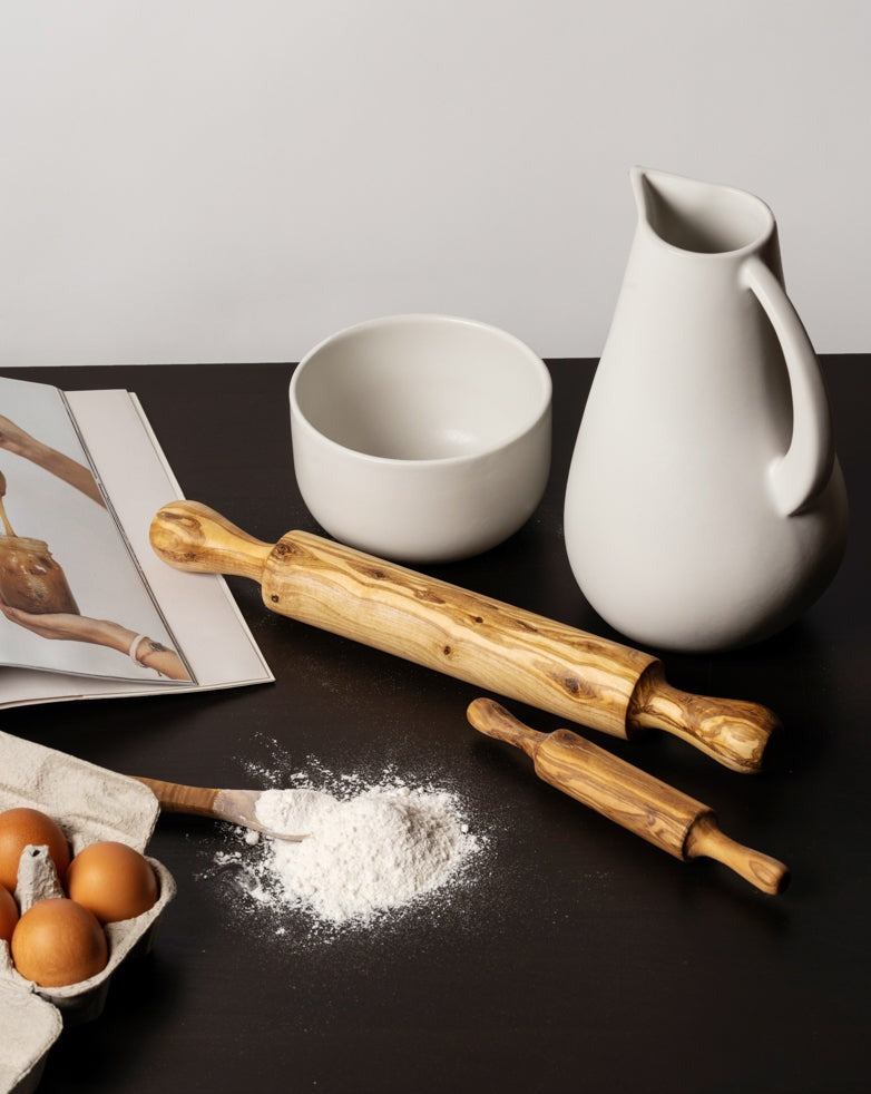 Two natural olive wood rolling pins placed on a dark surface, surrounded by baking elements including a pile of flour with a wooden spoon, brown eggs in a carton, an open cookbook, and minimalist white ceramics—a bowl and a pitcher—creating a warm, artisan kitchen scene