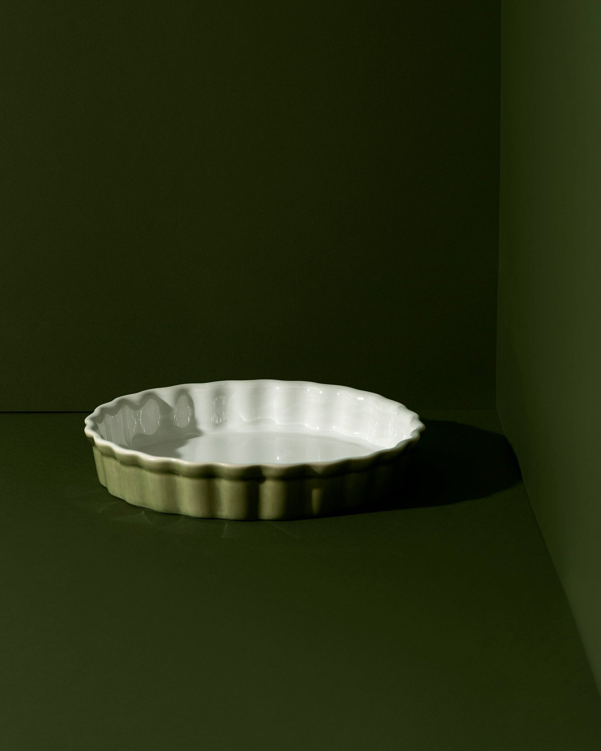 A white rounded stoneware pie dish with wavy side walls displayed on a neutral background, accompanied by a wooden cutting board with a pie on it, some garlic bulbs, and a small white bowl.
