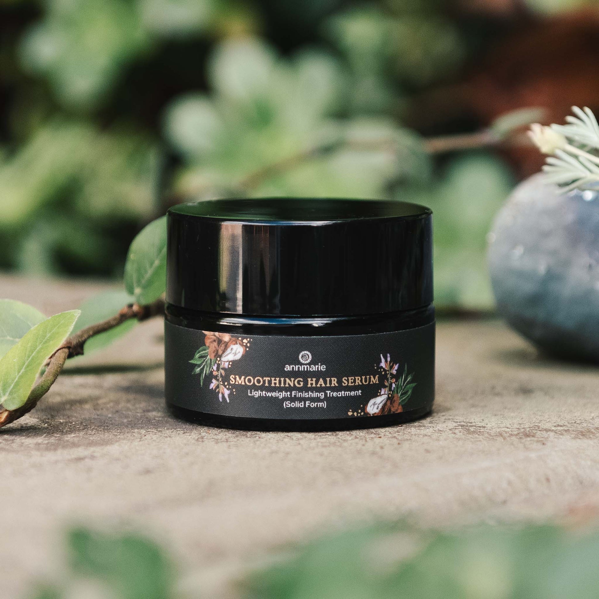 Smoothing Hair Serum lightweight finishing treatment in solid form displayed in a black jar on a natural surface with soft greenery in the background.