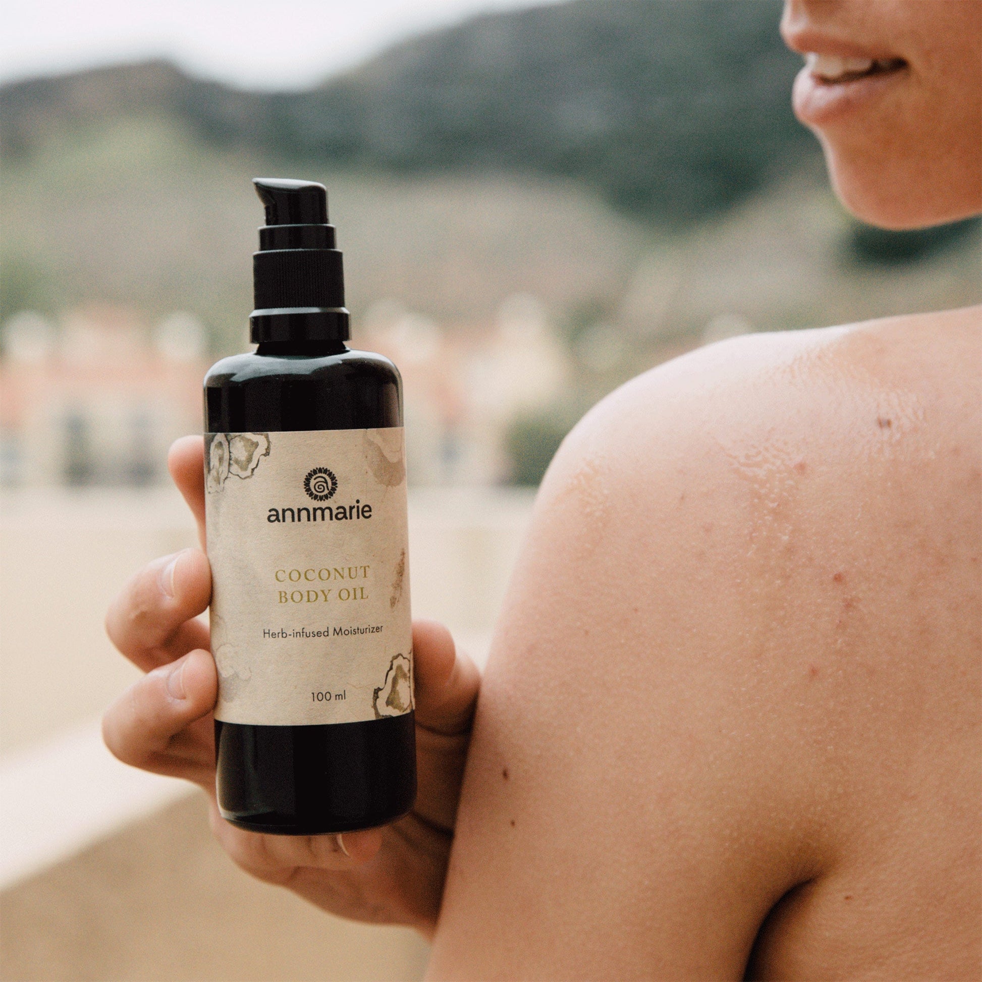 Coconut Body Oil herb-infused moisturizer in 100 ml pump bottle held beside glowing moisturized shoulder outdoors.