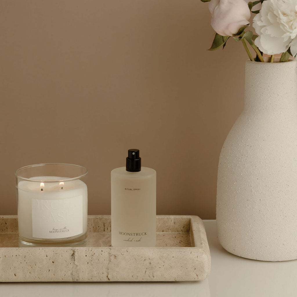 Candle, bottle, and vase with flowers on a marble tray against a beige wall.