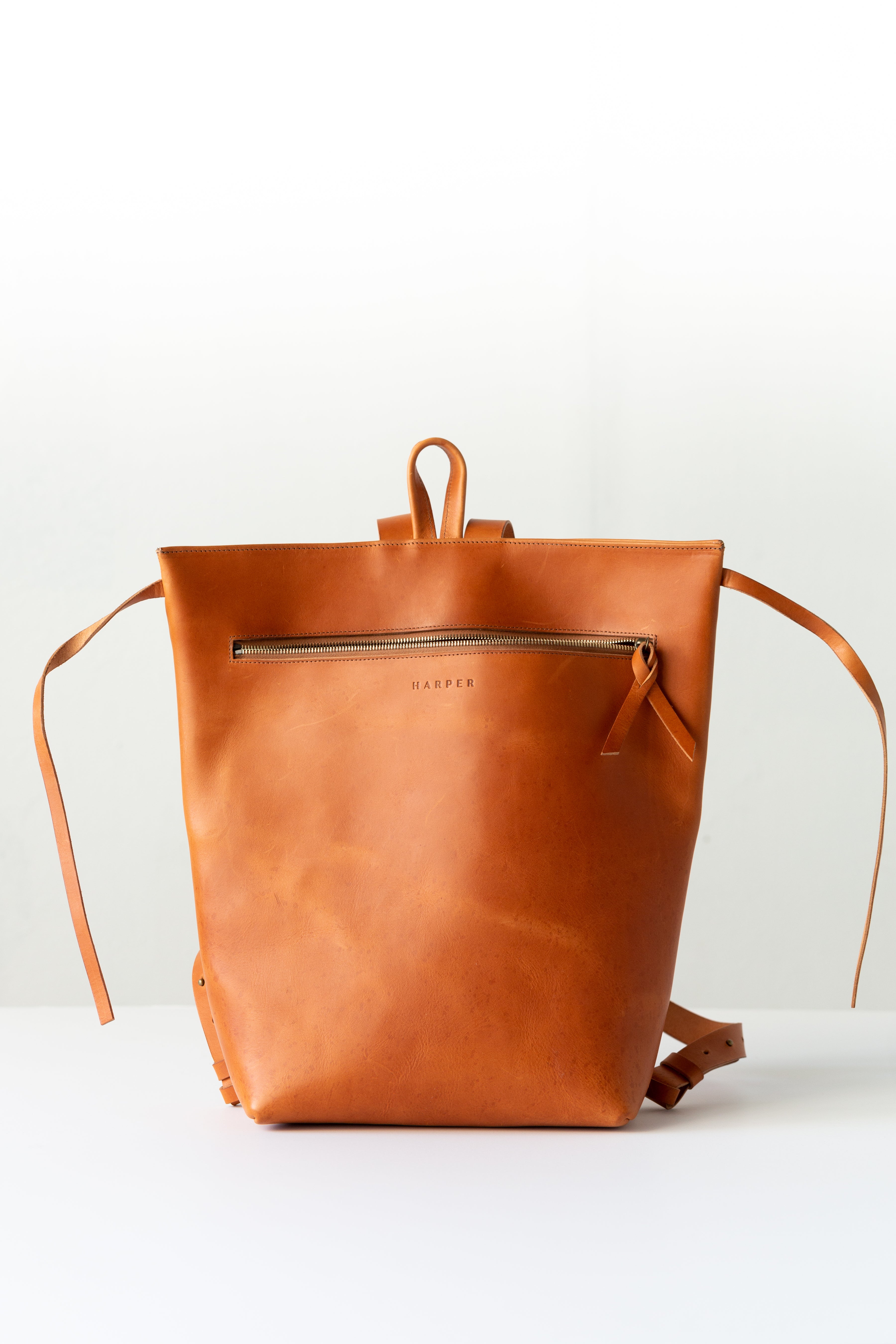 A woman holds out her arm, which is looped through the shoulder straps of a cognac brown leather backpack. The top corners of the backpack are cinched with straps that knot over a zipper, creating a unique shape.