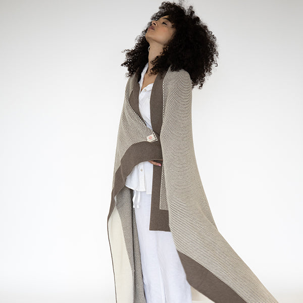 Woman wrapped in a beige organic cotton knit throw with a dark brown border over a white outfit.