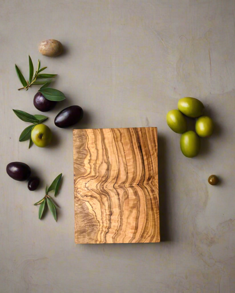 A rectangular olive wood chopping board placed on a gray surface, surrounded by assorted green and black olives, olive leaves, and a stone, creating a natural and rustic composition.
