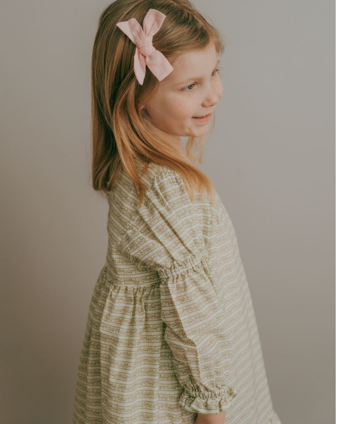 A child wearing a blush vine stripe dress with soft shirred sleeves and a ruffled hem.