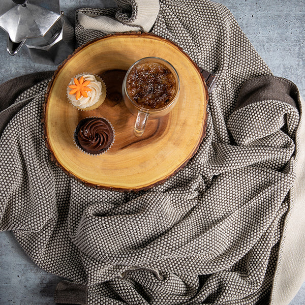 Wooden plate with a cup of coffee and  cupcake on a dark brown and white textured cotton blanket.