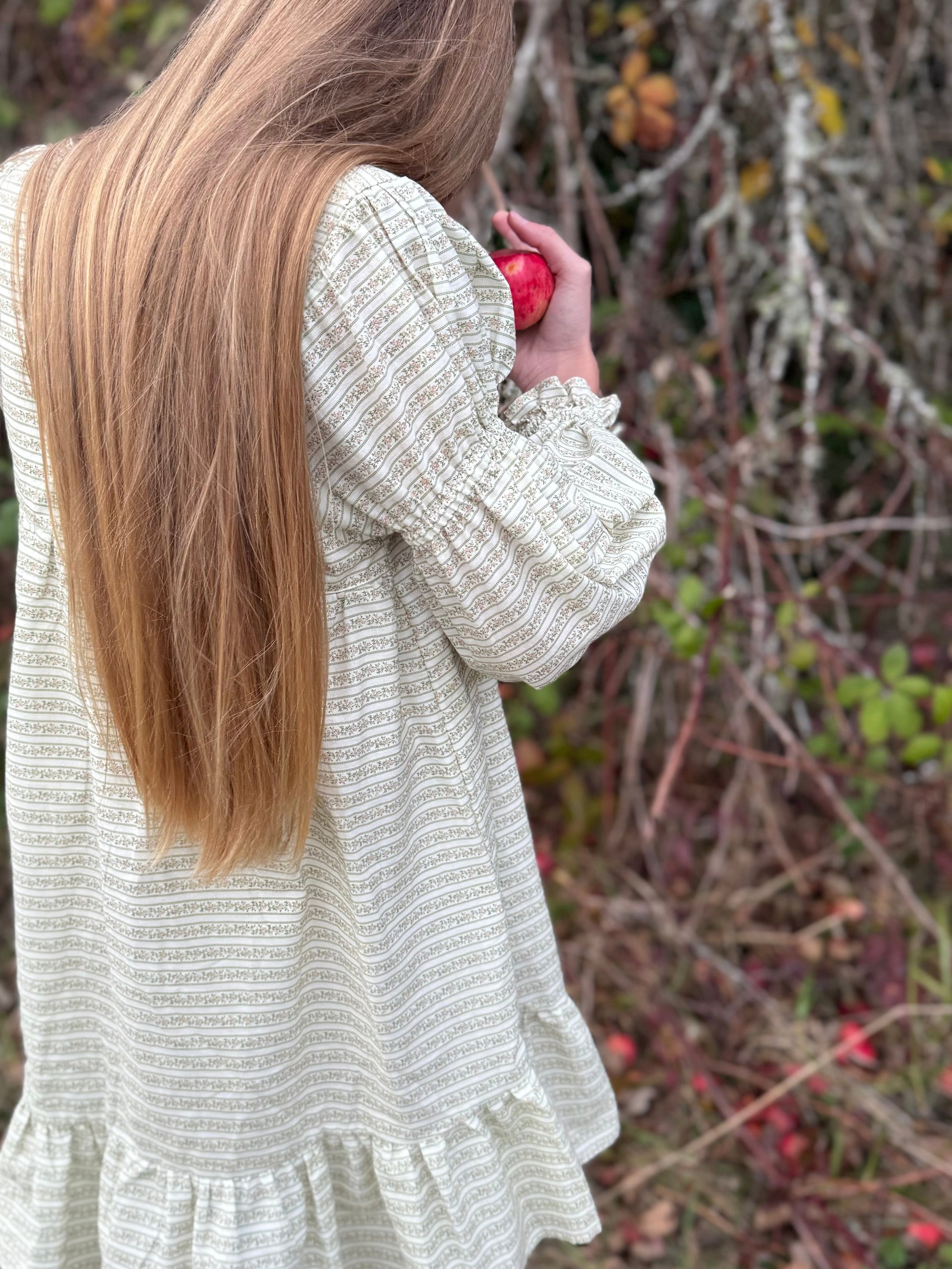 Person wearing a textured dress holding an apple in a natural setting with trees and berries.