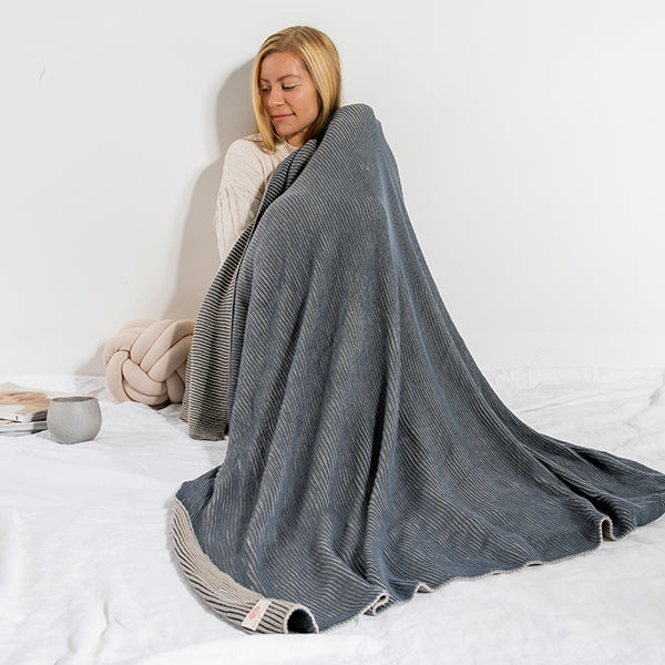 Woman wrapped in a blue and gray cotton throw blanket on a bed while laying next to a knotted cushion and enjoying a cup of coffee.
