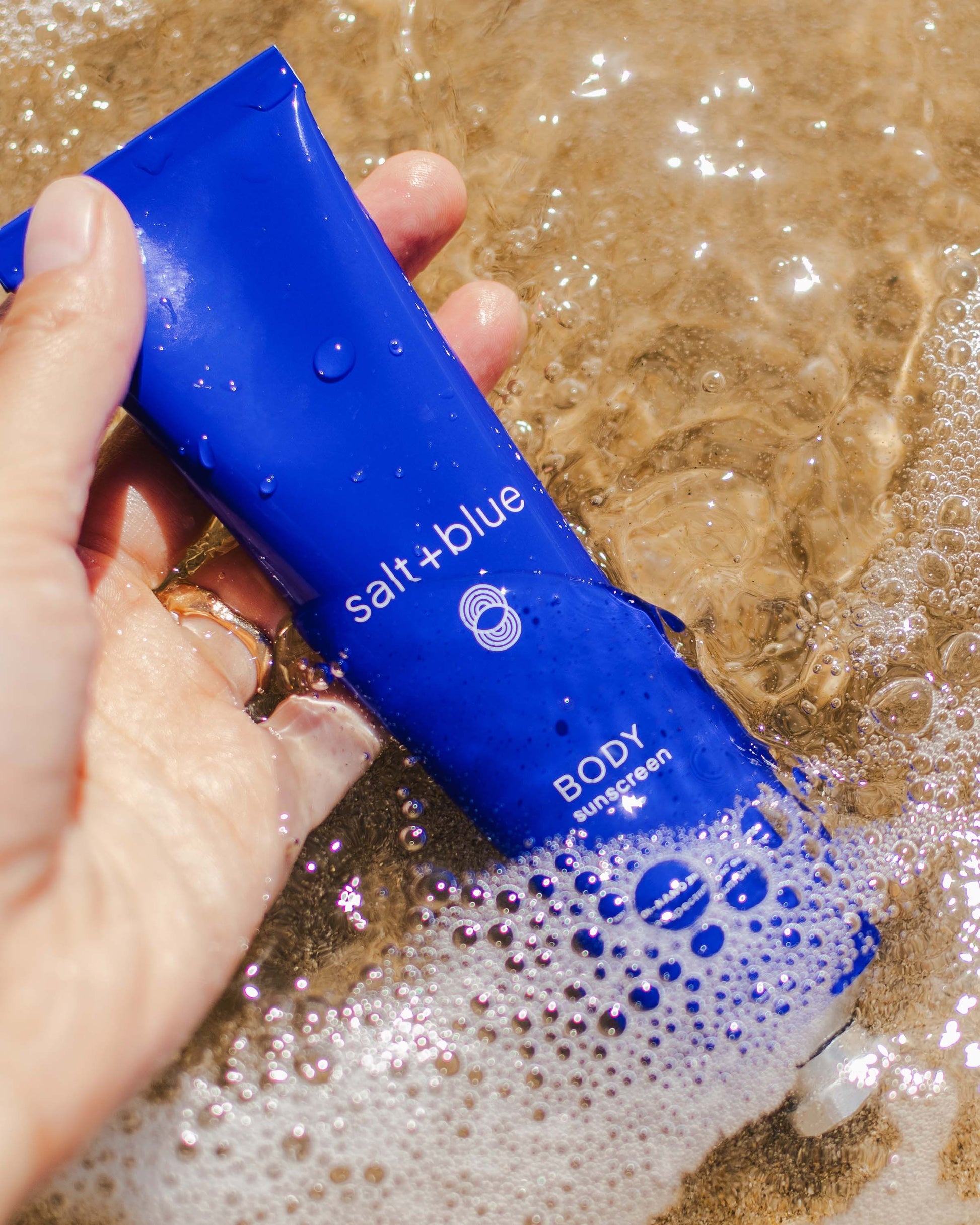 Blue sunscreen tube labeled 'salt+blue' being held over water with bubbles.