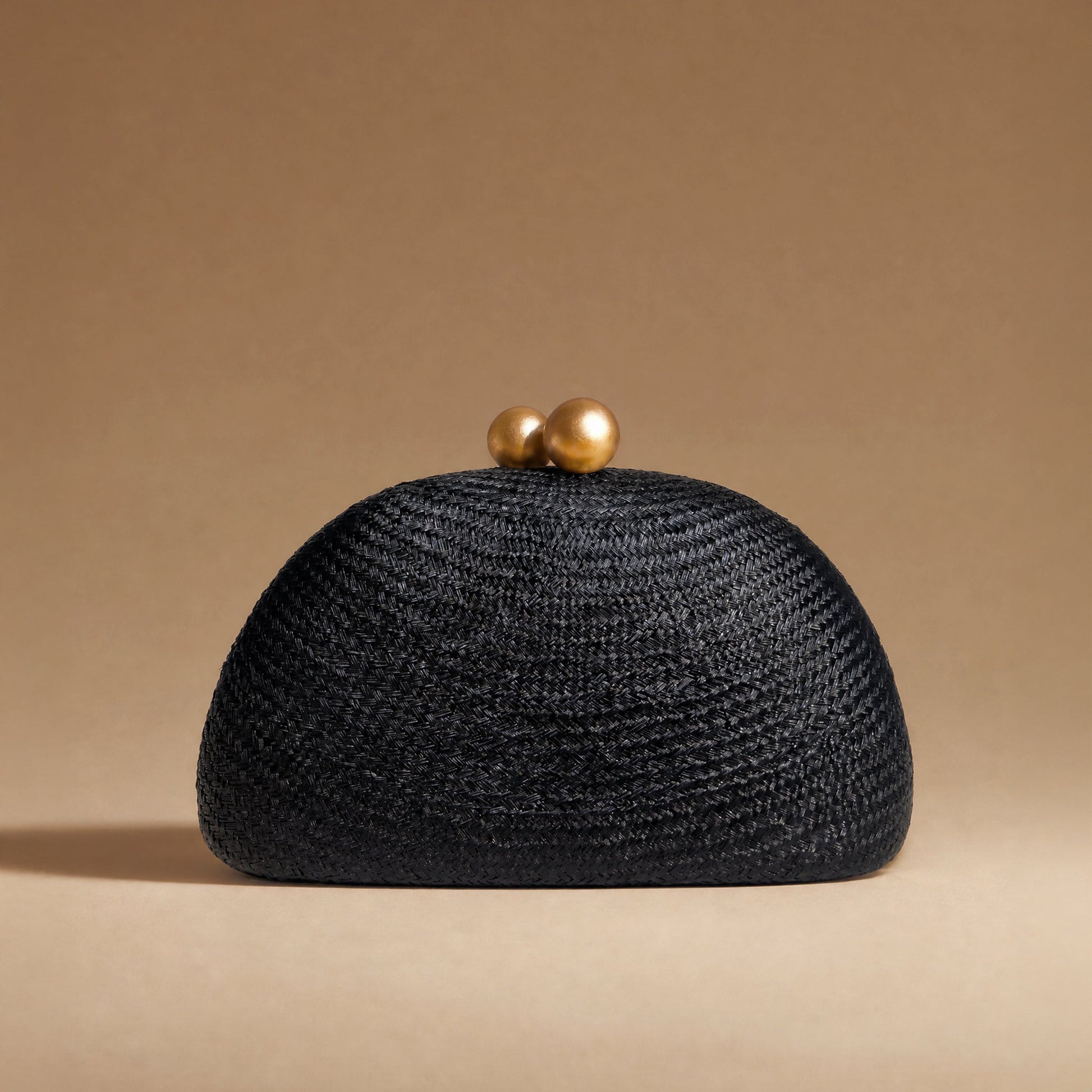 A black woven straw clutch purse with two gold-tone spherical clasp beads on top, set against a solid warm beige background.