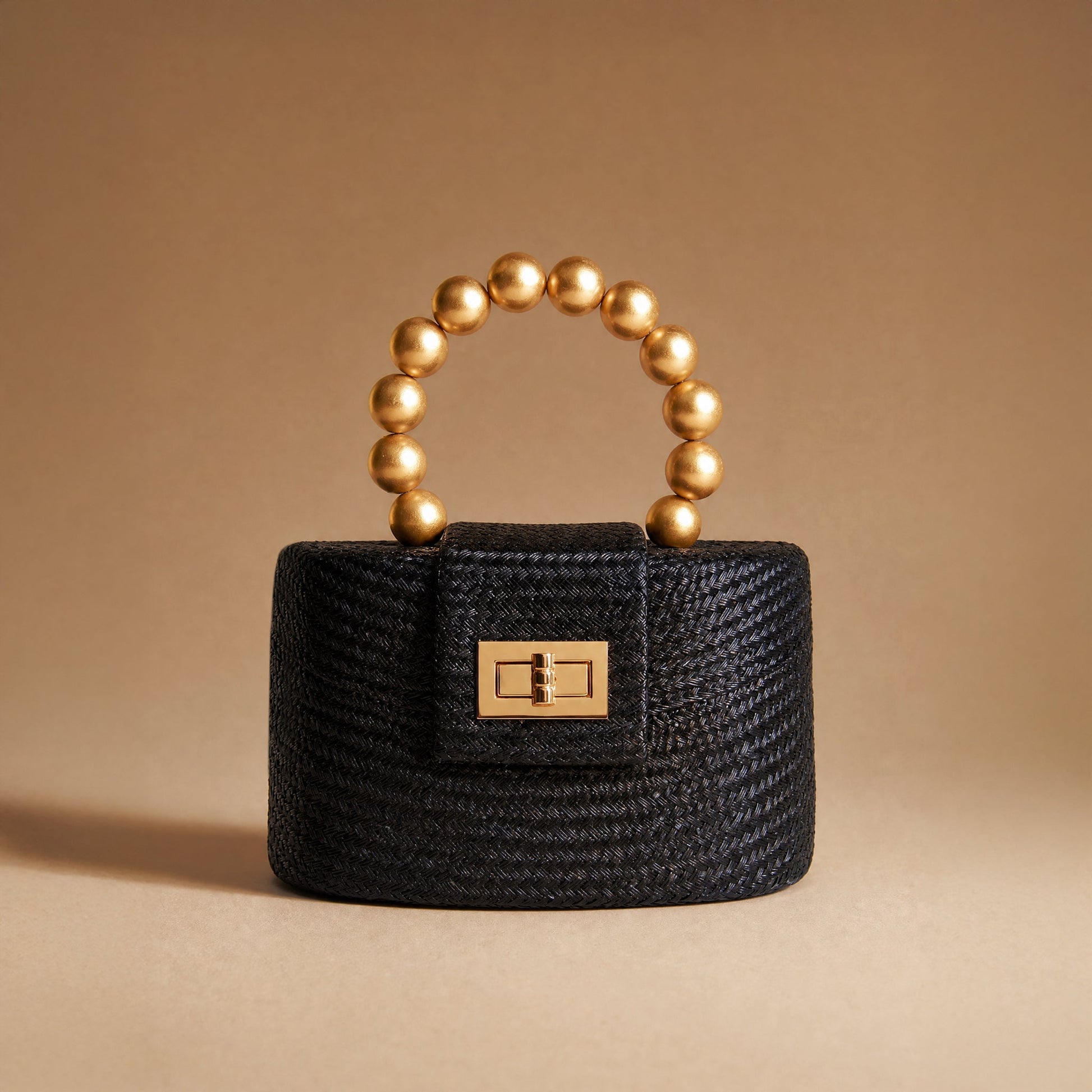 A small structured black woven handbag with a gold-tone clasp and a curved top handle made of metallic gold beads, photographed on a warm beige studio background.