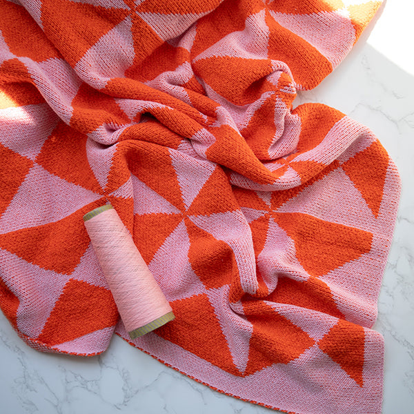 Pink and neon orange blanket with geometric designs with a spool of pink thread on a marble surface.