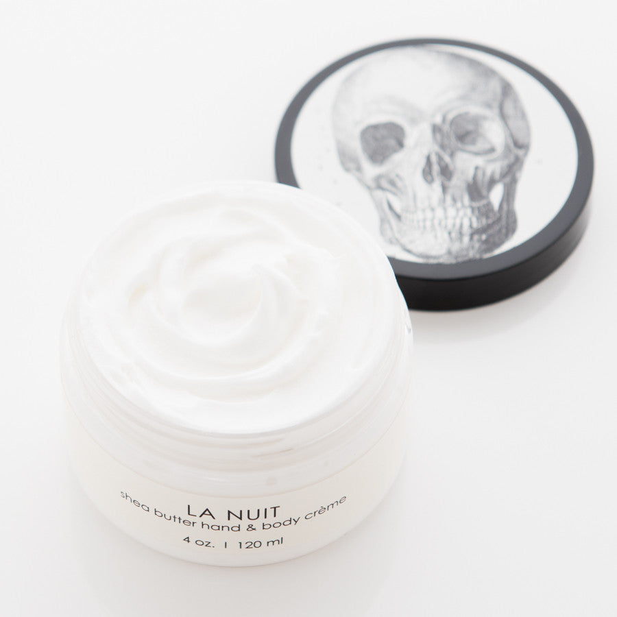 A container of La Nuit shea butter hand and body crème with the lid off. The lid, displayed next to the container, features a black and white skull illustration. The crème is white and appears smooth and creamy. The container holds 4 oz (120 ml) of product.