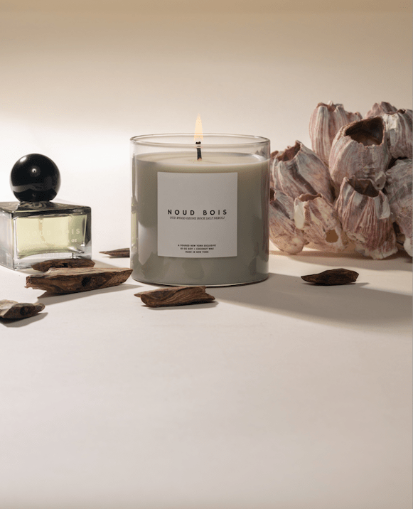 Candle labeled 'Noud Bois' with a oud and rocksalt arrangement and perfume bottle on a neutral background