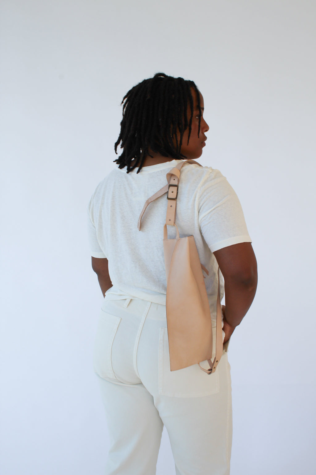 A woman wears a natural, undyed leather fanny pack shoulder bag against a white background. Her back faces the camera and the bag rests against her back.