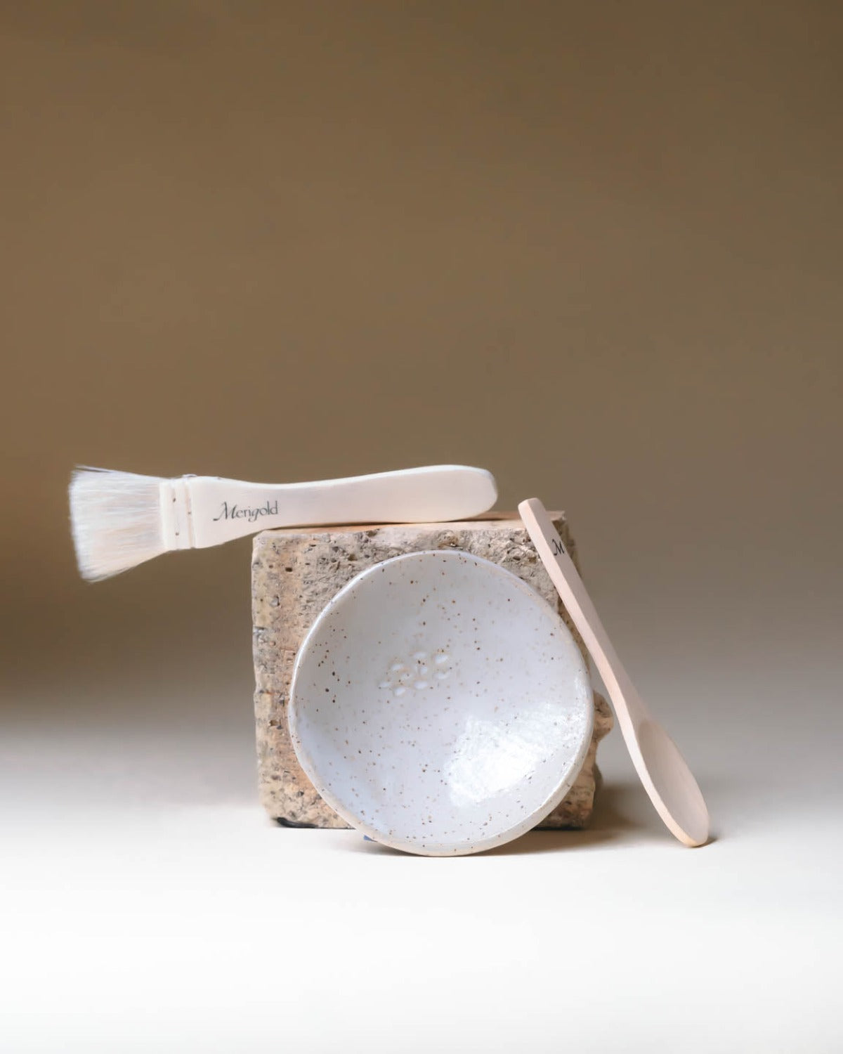 Merigold masking ritual tools—ceramic bowl, wooden spoon, and mask brush—styled on natural stone for clean skincare rituals.