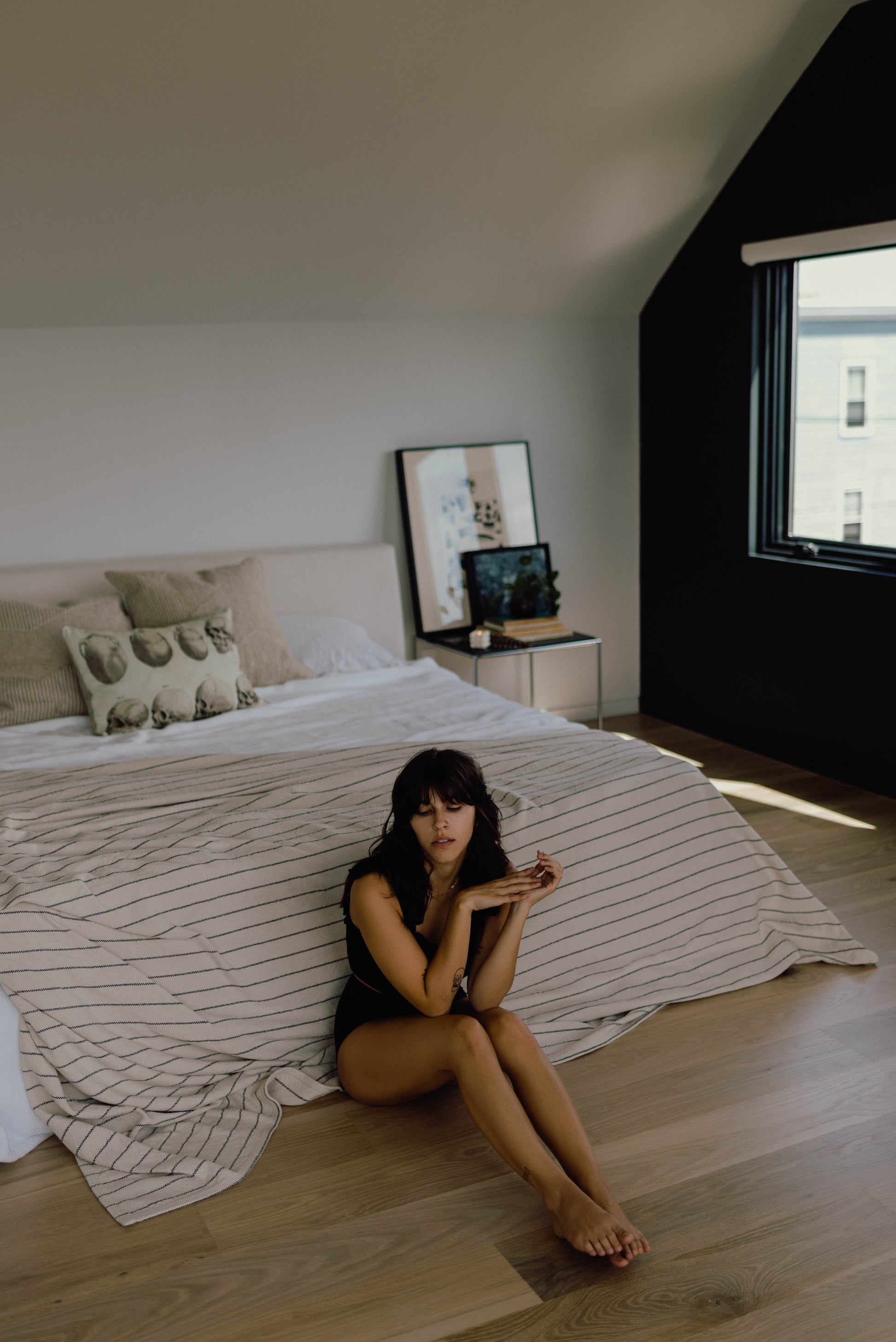 A luxurious bedroom scene showcasing a woman gracefully seated on a bed adorned with Evangeline linens. The pinstripe pattern adds elegance to this 100% Cotton bed blanket, available in king, queen, and twin sizes. Crafted with durability and washability in mind, this heirloom piece exudes timeless luxury.