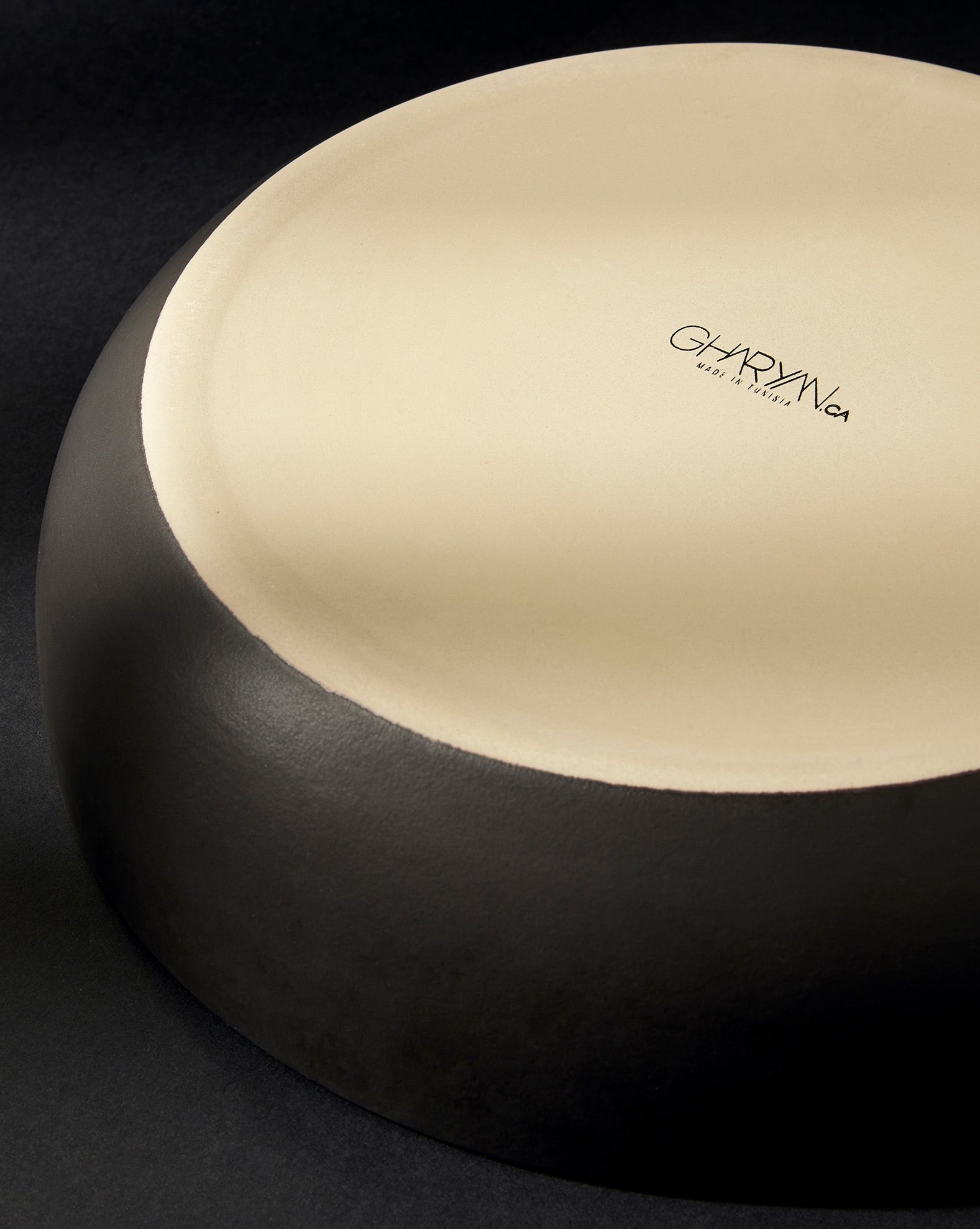 A matte black stoneware shallow serving bowl placed upside down against a dark background, with the “GHARYAN” logo visible on the base.