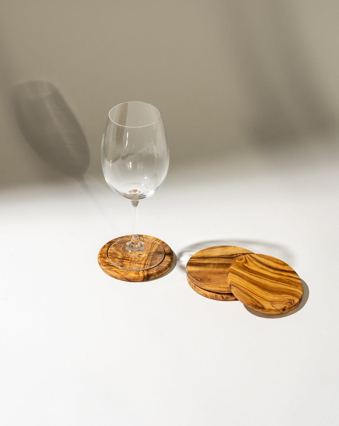 Three round olive wood coasters with natural grain patterns displayed on a white background, with a glass placed on one of them, highlighting their functional and aesthetic appeal.
