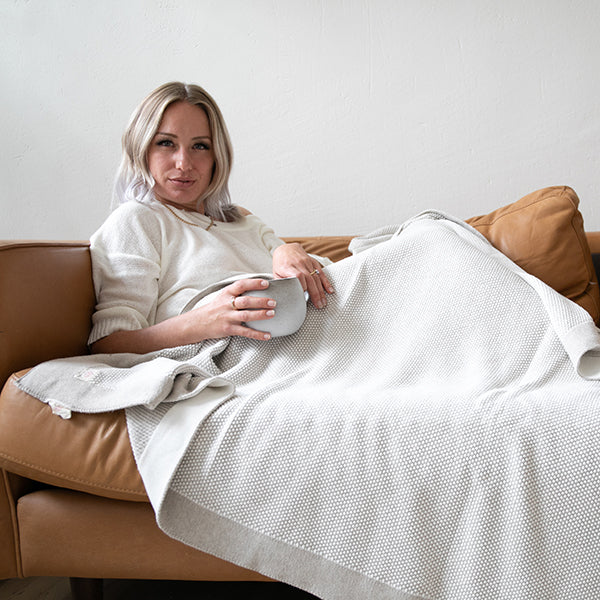 Woman sitting on a brown leather couch with a textured white cotton throw blanket with a light grey border.