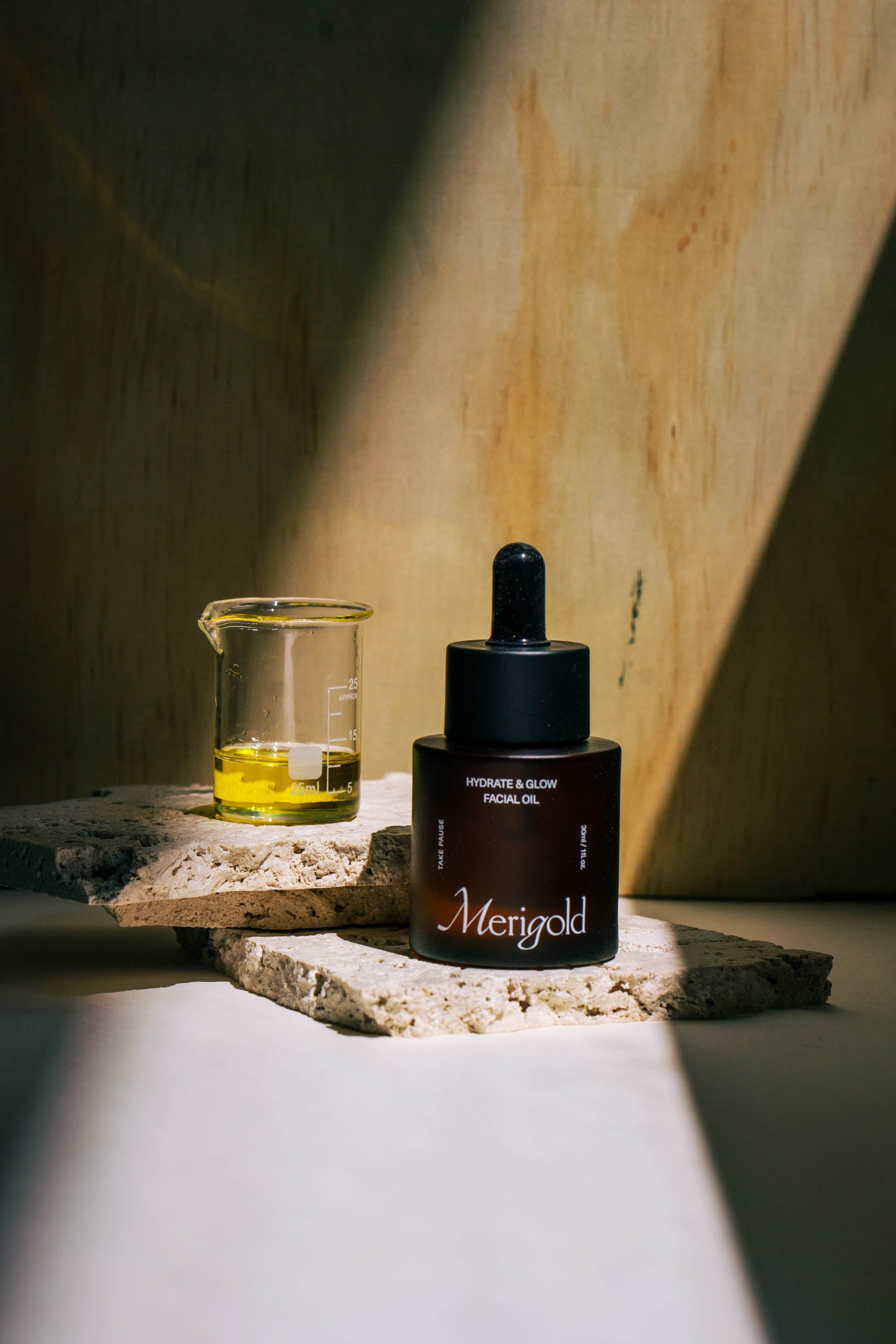 Merigold Hydrate & Glow Facial Oil with botanical ingredients in beaker, on natural stone — clean beauty face oil for radiant skin