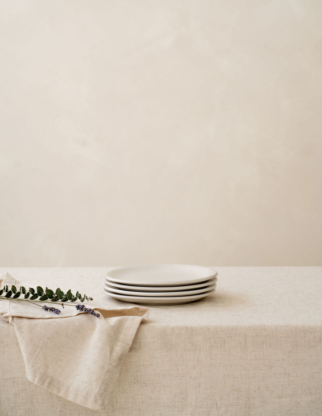 A set of white ceramic plates stacked on top of each other on a beige linen tablecloth with a sprig of herbs to the side and a napkin partially visible.