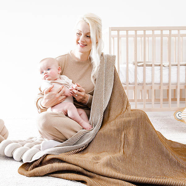 Woman holding an infant while wrapped in a copper coloured ribbed cotton blanket near a wooden crib. 