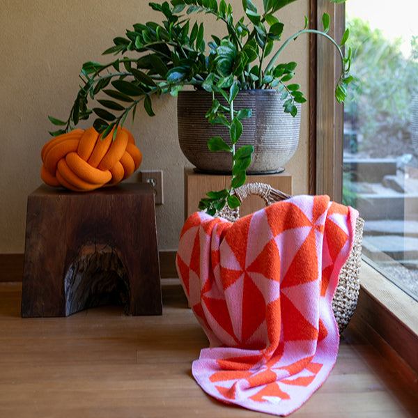 Orange knot pillow and geometric patterned neon orange and pink blanket on a wooden surface with plants in the background.