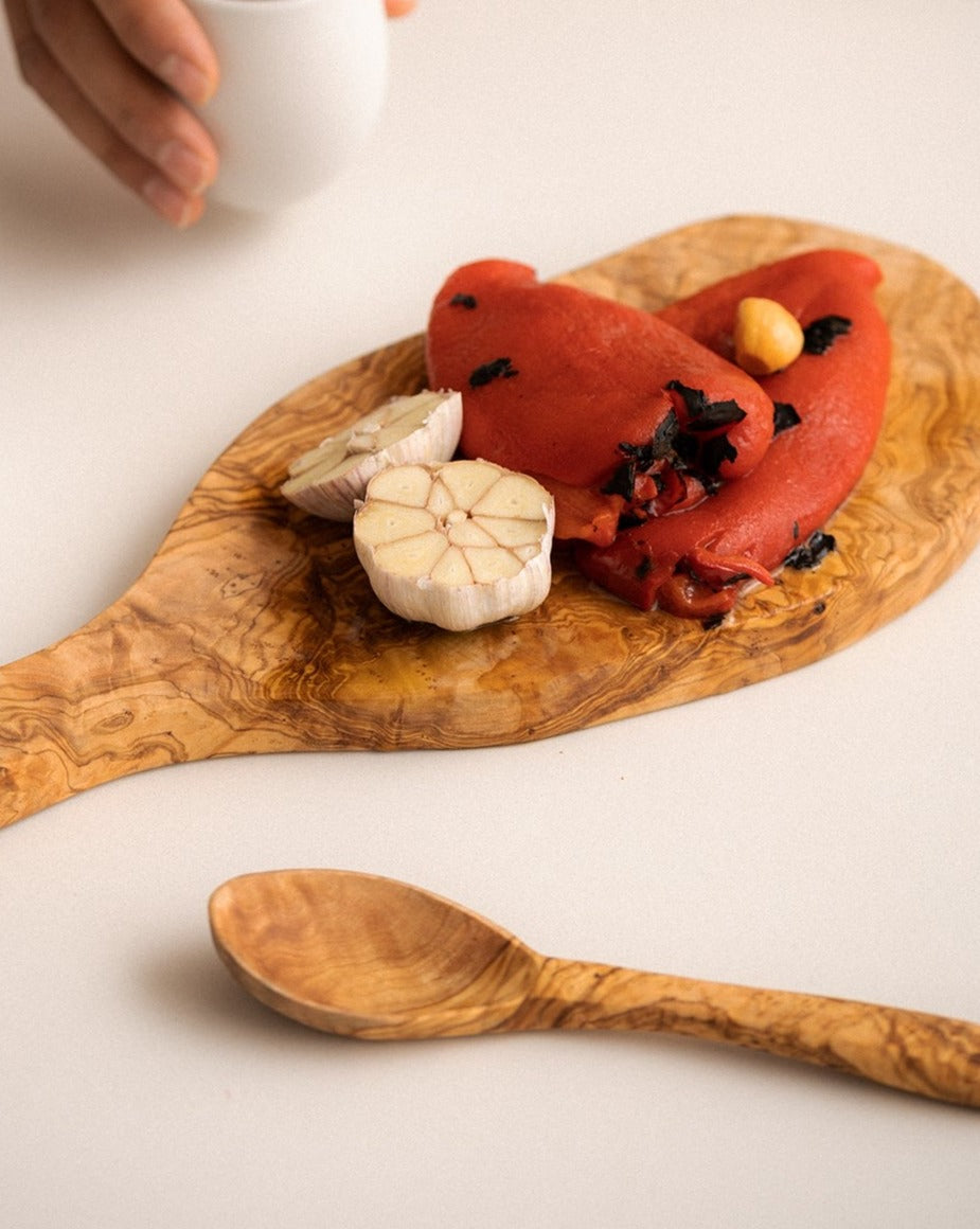 A natural olive wood kitchen spoon placed beside a wooden cheese board topped with various food items, all set against a dark background for a warm, rustic presentation.


