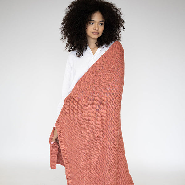 Woman wearing a rosewood pink knitted cotton blanket over her shoulders against a white background.