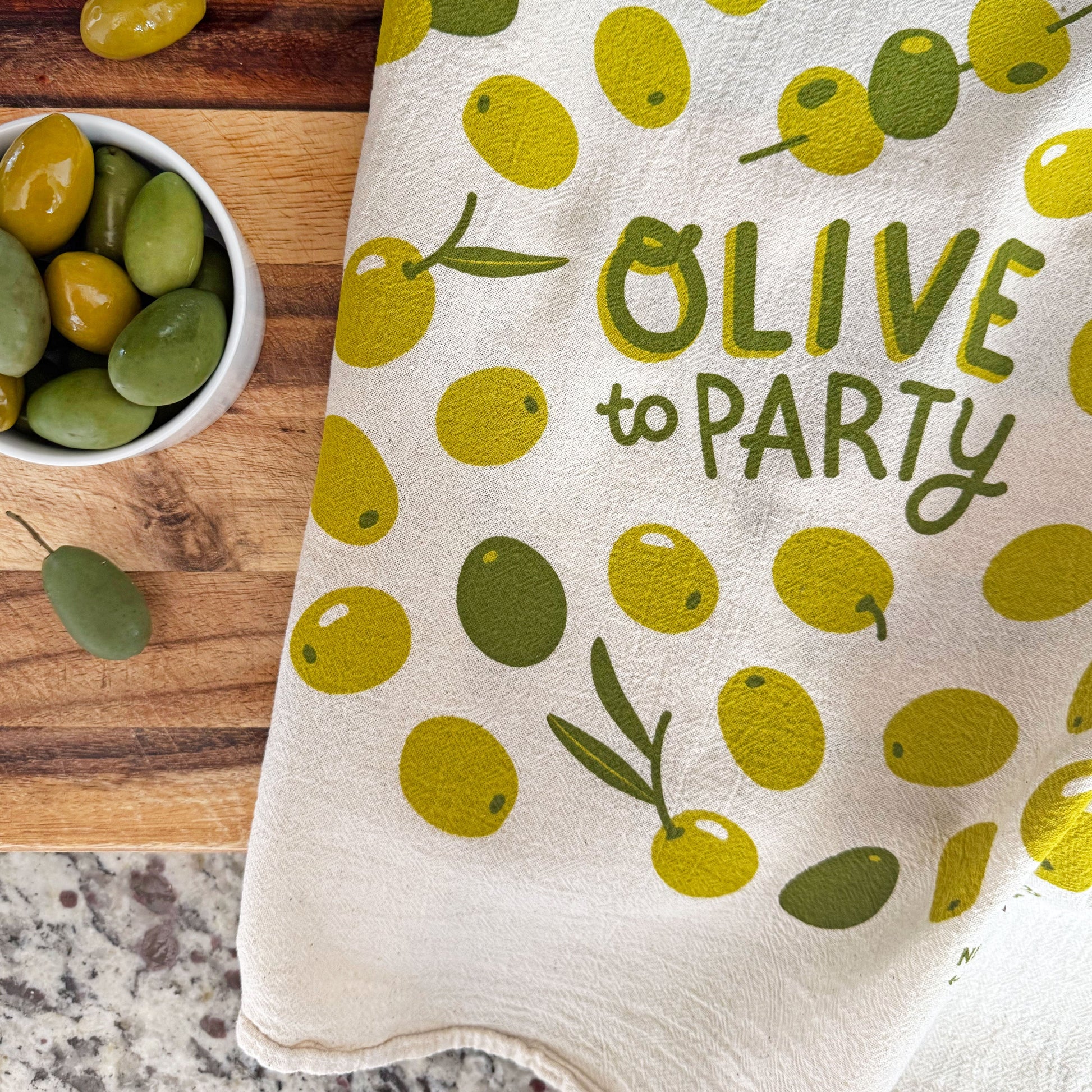 The Neighborgoods Olive dish towel featuring a olive illustration, laid next to bowl of green olives.