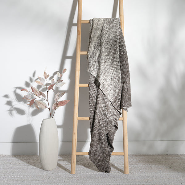 Dark brown and ombre textured throw draped over a wooden ladder against a white wall with a vase of dried flowers beside it.